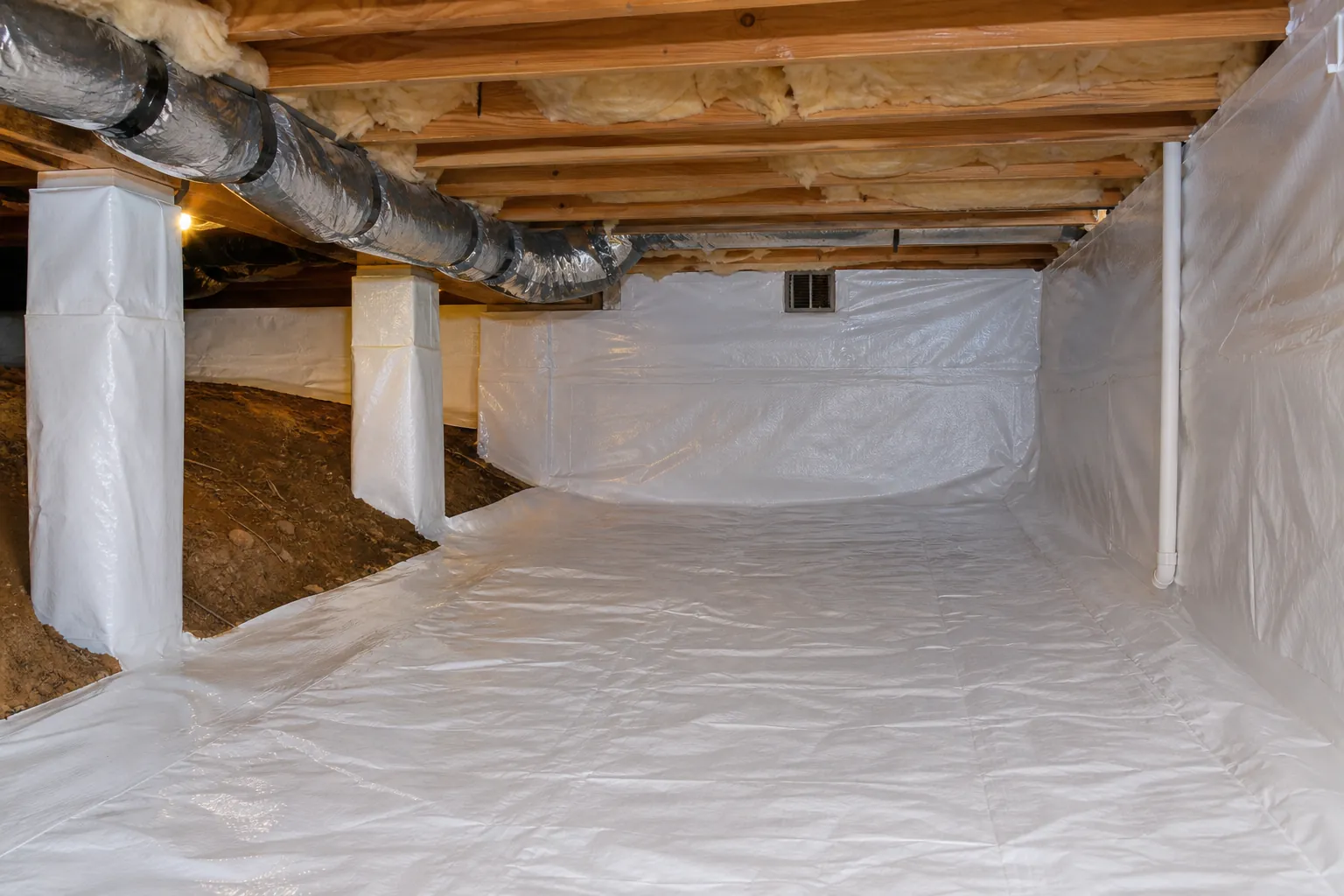 Crawl space with white vapor barrier covering the floor, walls and support piers beside exposed sloped dirt under wooden joists.