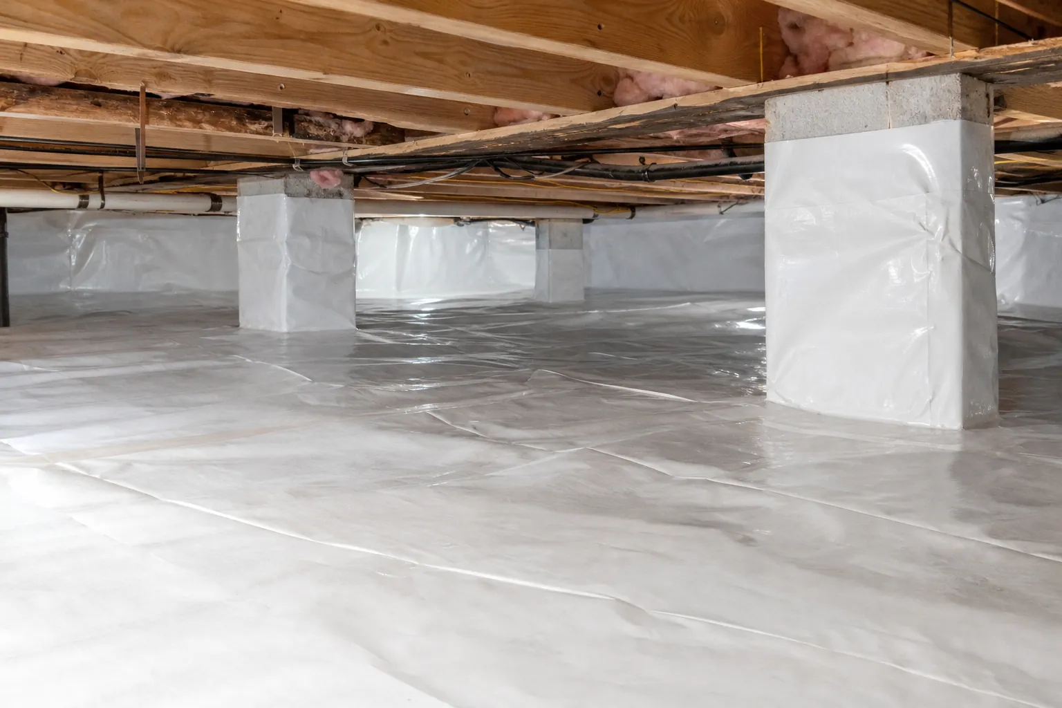 White vapor barrier covers a crawl space floor, walls, and support piers beneath exposed wooden joists and pipes.