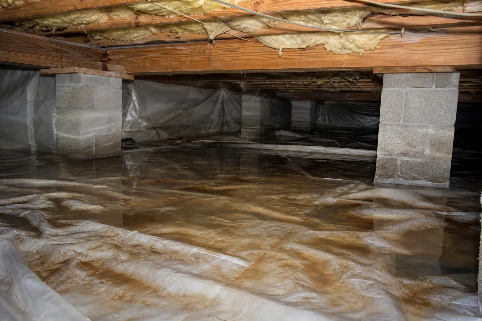 Standing water covers wrinkled plastic sheeting in a crawl space beneath wooden joists, exposed insulation, and concrete block piers.
