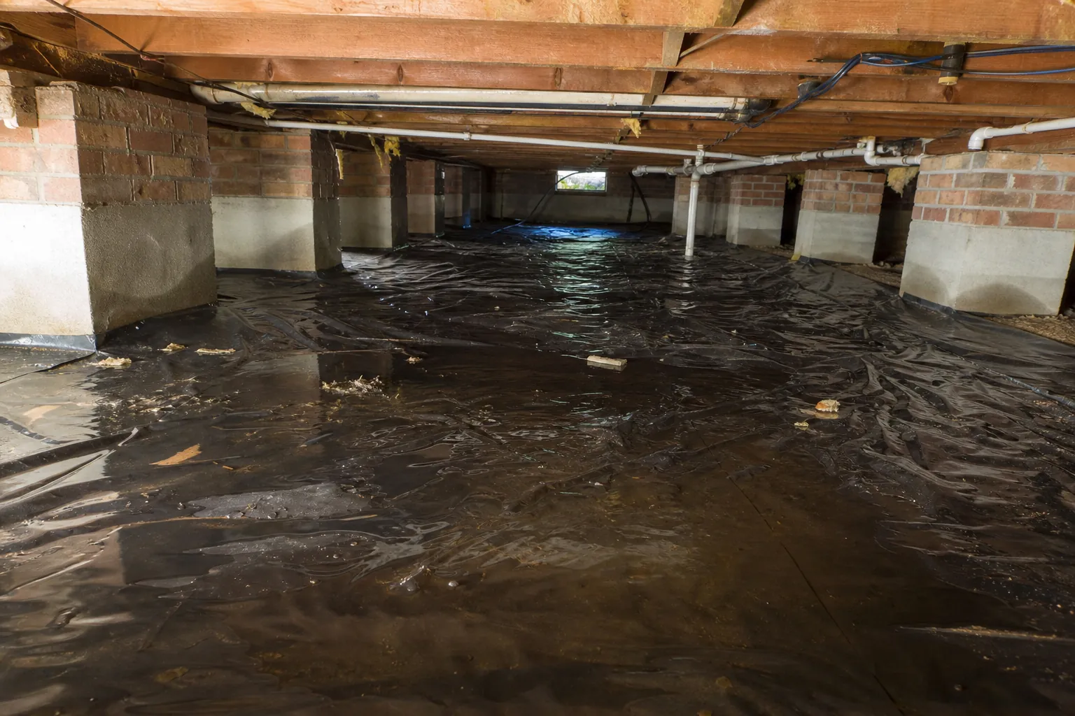 Black plastic vapor barrier covers a crawl space floor beneath exposed wooden joists, pipes, and brick support piers.