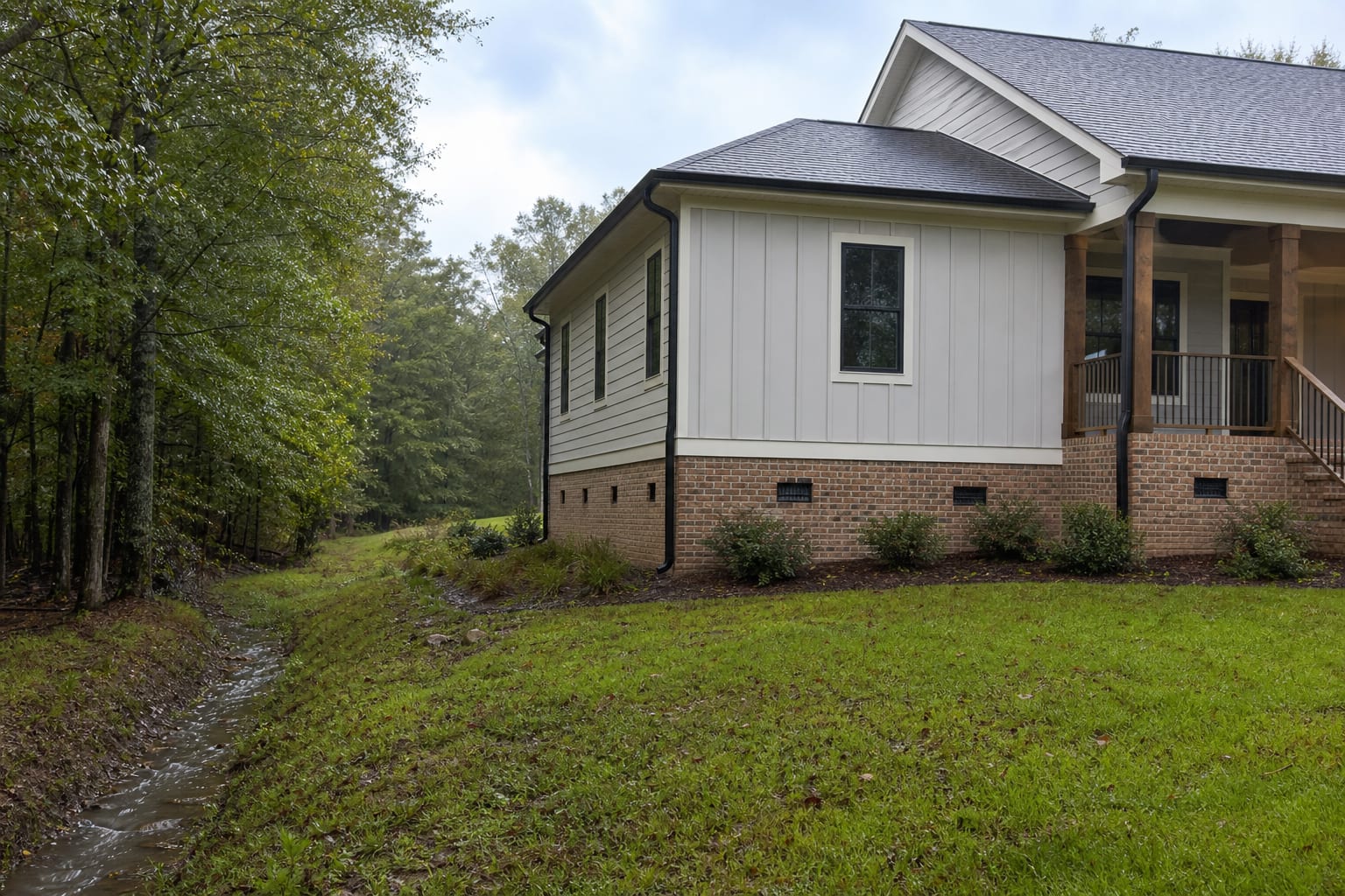 Rural-edge home with crawl space vents and a wet drainage swale near a wooded boundary